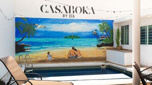 Een muurschildering van een strandscène met palmbomen en een stel op het strand, met de tekst 'CASABOKA BY ECA'.