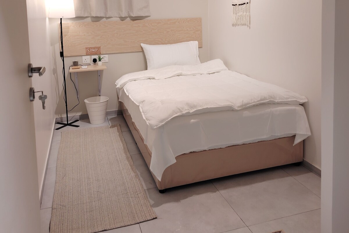 A minimalist bedroom with a bed, bedside table, lamp, and a wall-mounted artwork.
