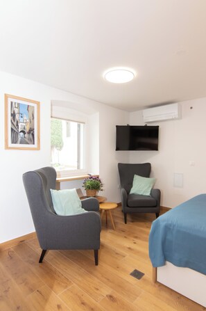 Une chambre moderne avec deux fauteuils gris, une petite table en bois ornée d’une plante, un lit recouvert d’une couverture bleue, une fenêtre donnant sur des immeubles et un téléviseur fixé au mur.