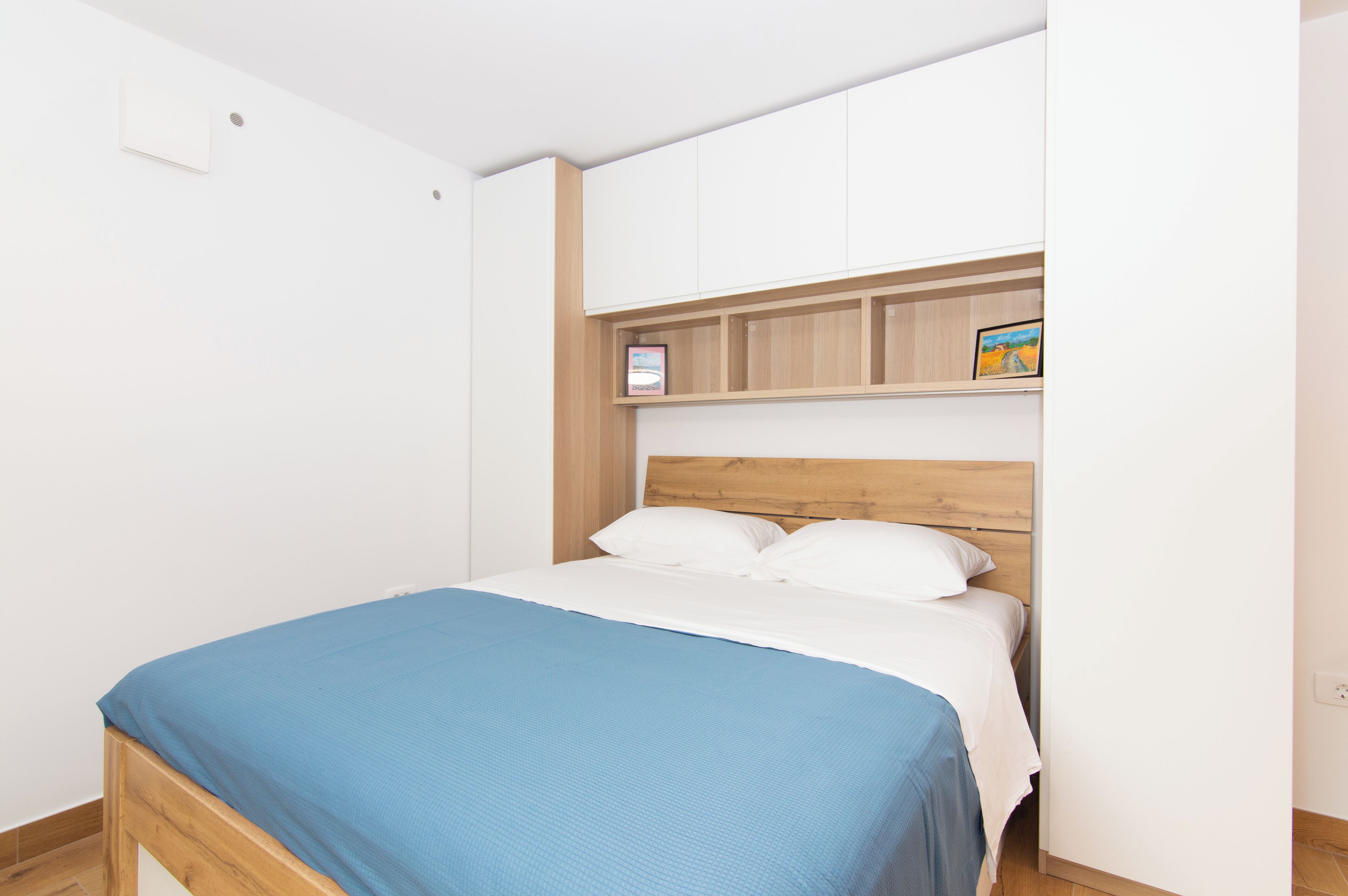 A bedroom with a wooden bed frame, white bedding, and a blue blanket. There is a shelf above the bed with a framed picture.