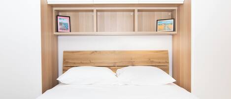 A bed with a wooden headboard, white bedding, and a blue blanket. Above the bed, there are two wooden shelves with framed pictures.