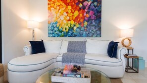 A modern living room with a white sofa, two side lamps, a colorful abstract painting, and a glass coffee table with books. - Elegant 3 BR Ground-Floor Apartment with Hot Tub (Cocle)