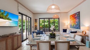 A dining area with a glass table, chairs, and a view of a tropical garden through large windows.