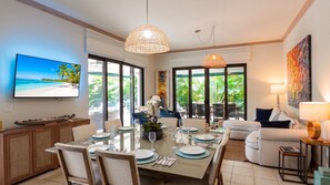 Un espace de repas moderne, avec une grande table dressée pour un repas, un téléviseur fixé au mur et une vue sur une végétation luxuriante à l’extérieur.