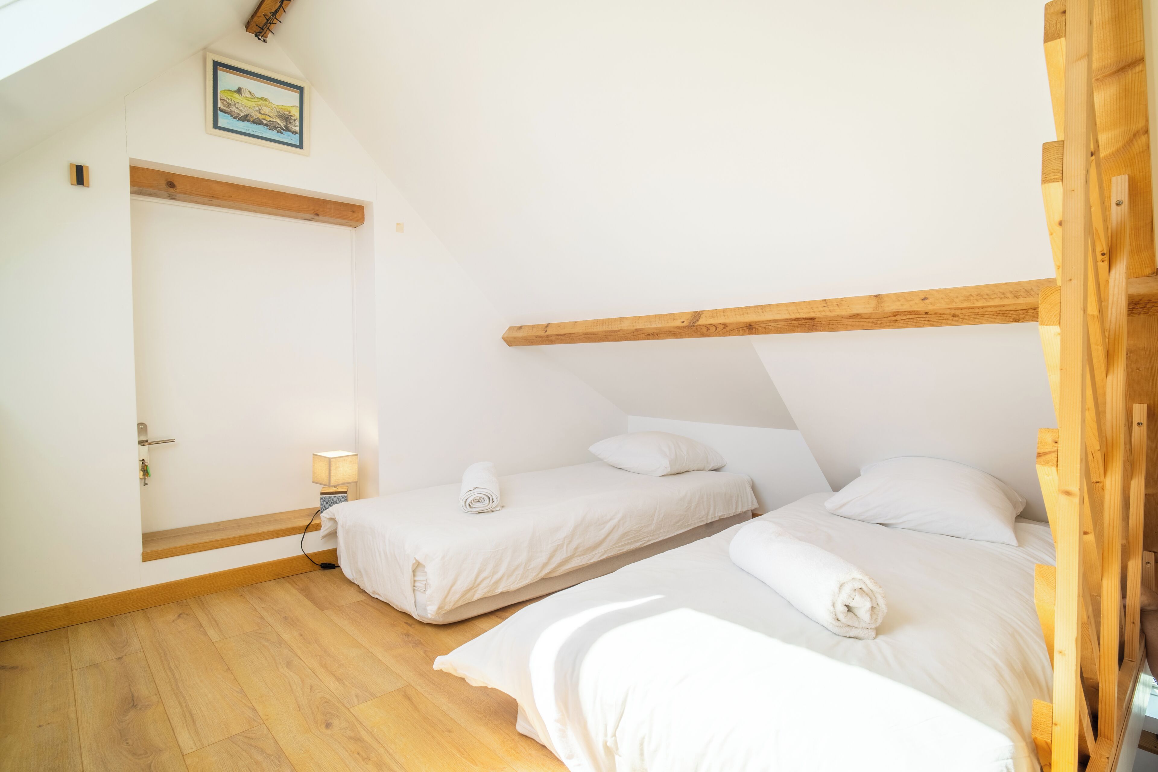 A bedroom with two beds, wooden beams, a door, and a small bedside table with a lamp.