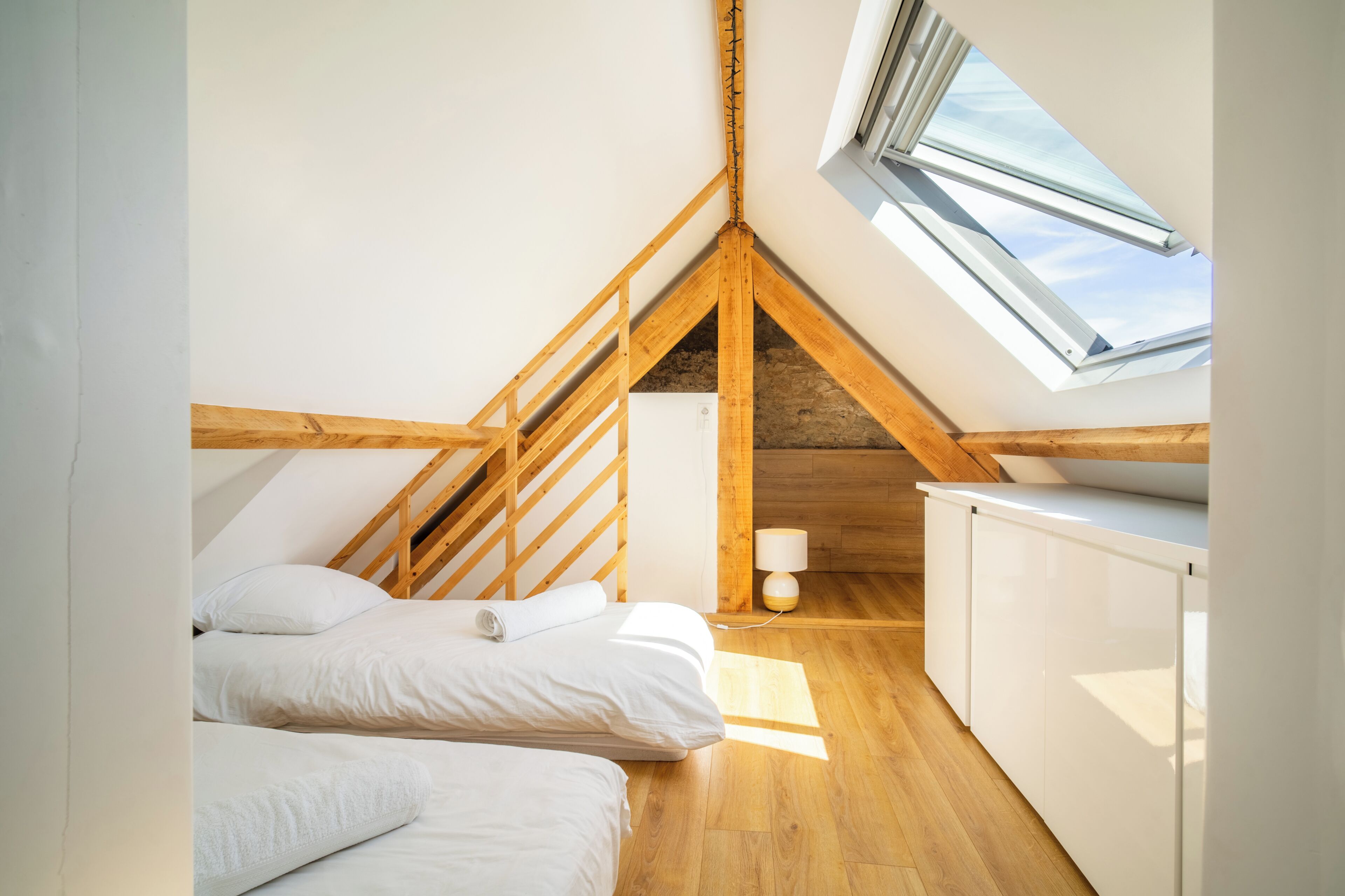 Un dormitorio con techo inclinado, vigas de madera, tragaluz, cama con ropa blanca y mesita de noche con lámpara.