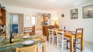 A dining area with a wooden table set for two, a kitchen in the background, and a framed seascape on the wall. - 3 *** village house, bright veranda opening onto terrace and large garden (Quiberon)