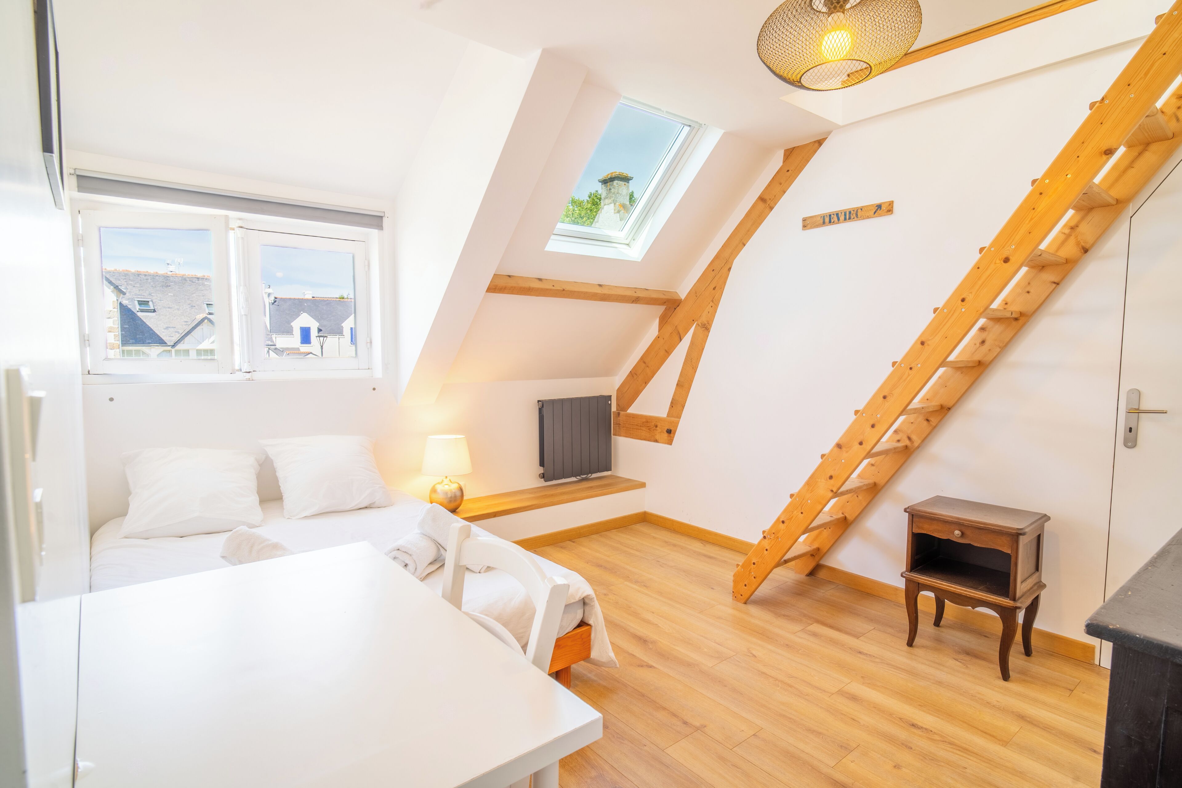 Una camera da letto con una zona notte soppalcata, una scala in legno, un tavolino e una finestra che si affaccia su edifici.
