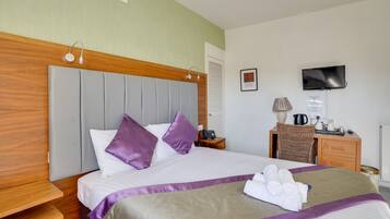 Habitación de hotel con cama, cabecera de madera, televisor montado en la pared, escritorio con silla y mesita de noche con lámpara y teléfono.