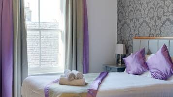 A hotel room with two beds, a window with curtains, a nightstand with a lamp, and a patterned wallpaper.