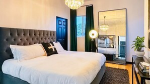 A bedroom with a large bed, a dark headboard, a chandelier, a mirror, and a bedside table with a lamp.