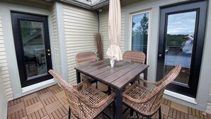 An outdoor patio with a wooden table and wicker chairs, a beige umbrella, and a view of a residential building with a garage door and a window.