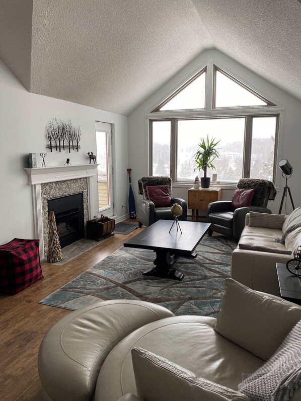 A spacious living room with a fireplace, leather sofas, a coffee table, and a large window offering a view of a snowy landscape.
