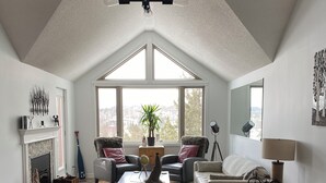 A spacious living room with a high ceiling, a fireplace, a dining table, and a large window offering a view of the outdoors.