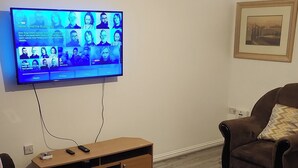 Una sala de estar con un televisor de pantalla plana montado en la pared, un mueble de televisión de madera, dos sillones marrones, una alfombra gris y un cuadro enmarcado en la pared.