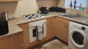 Cocina compacta con armarios de madera, estufa a gas, horno empotrado, lavadora y una ventana con persianas.
