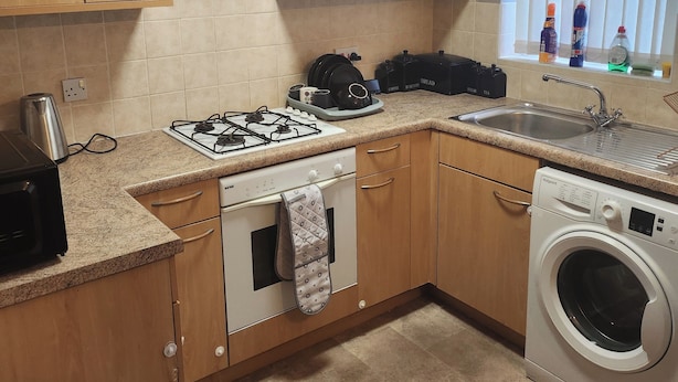 A compact kitchen with wooden cabinets, a gas stove, a built-in oven, a washing machine, and a window with blinds. - City Centre - 2 Bedrooms - Pet Friendly - Garden (Manchester)