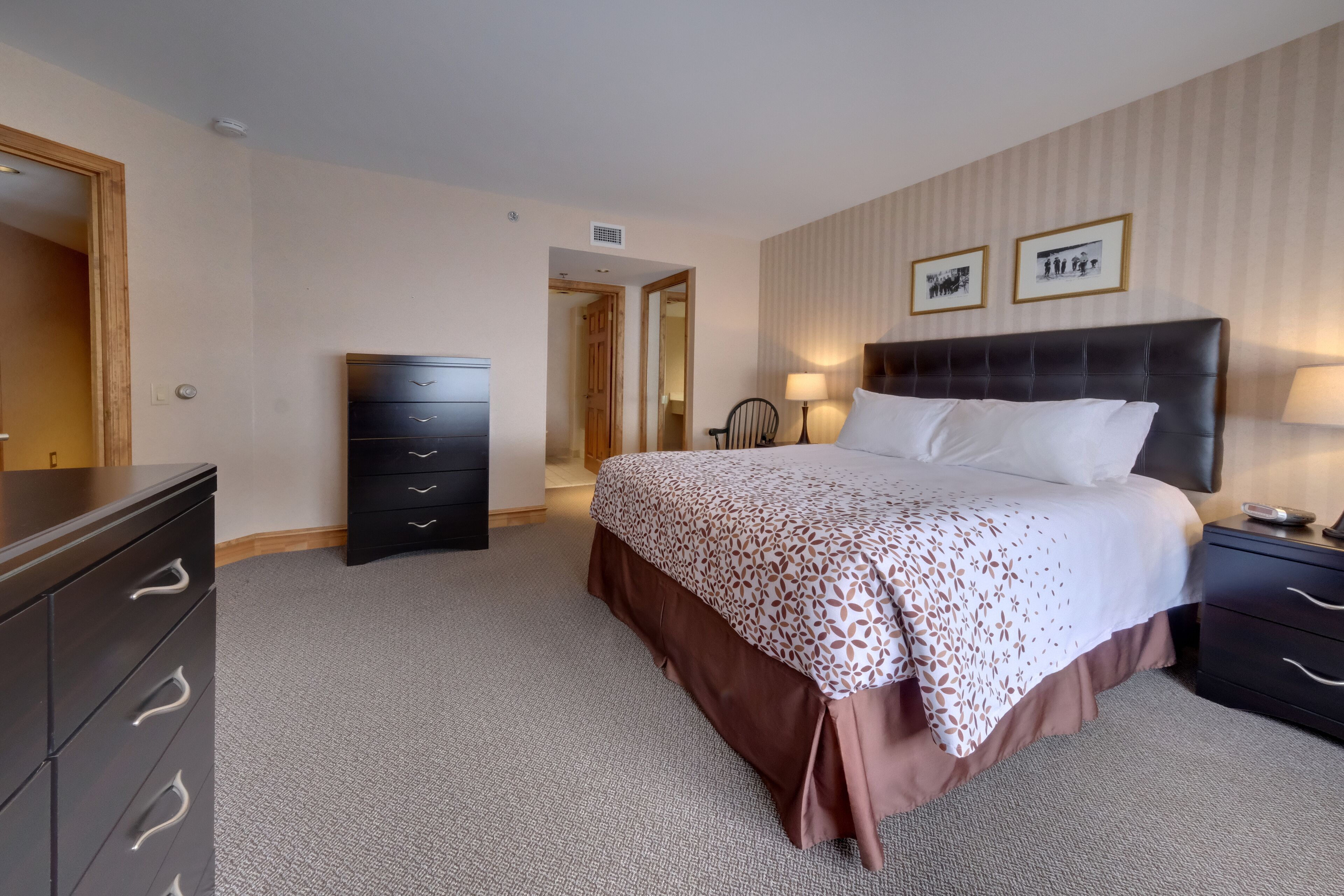 Habitación de hotel con una cama grande, una cómoda, una mesita de noche y una puerta que da a otra habitación.