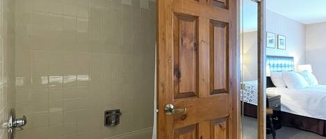 A bathroom with a glass-enclosed shower, a wooden door, and a visible bedroom with a bed and a nightstand.