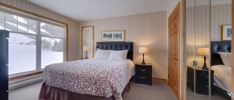 A hotel room with a large bed, two bedside tables with lamps, a wardrobe, and a large window with a view of a snowy landscape.