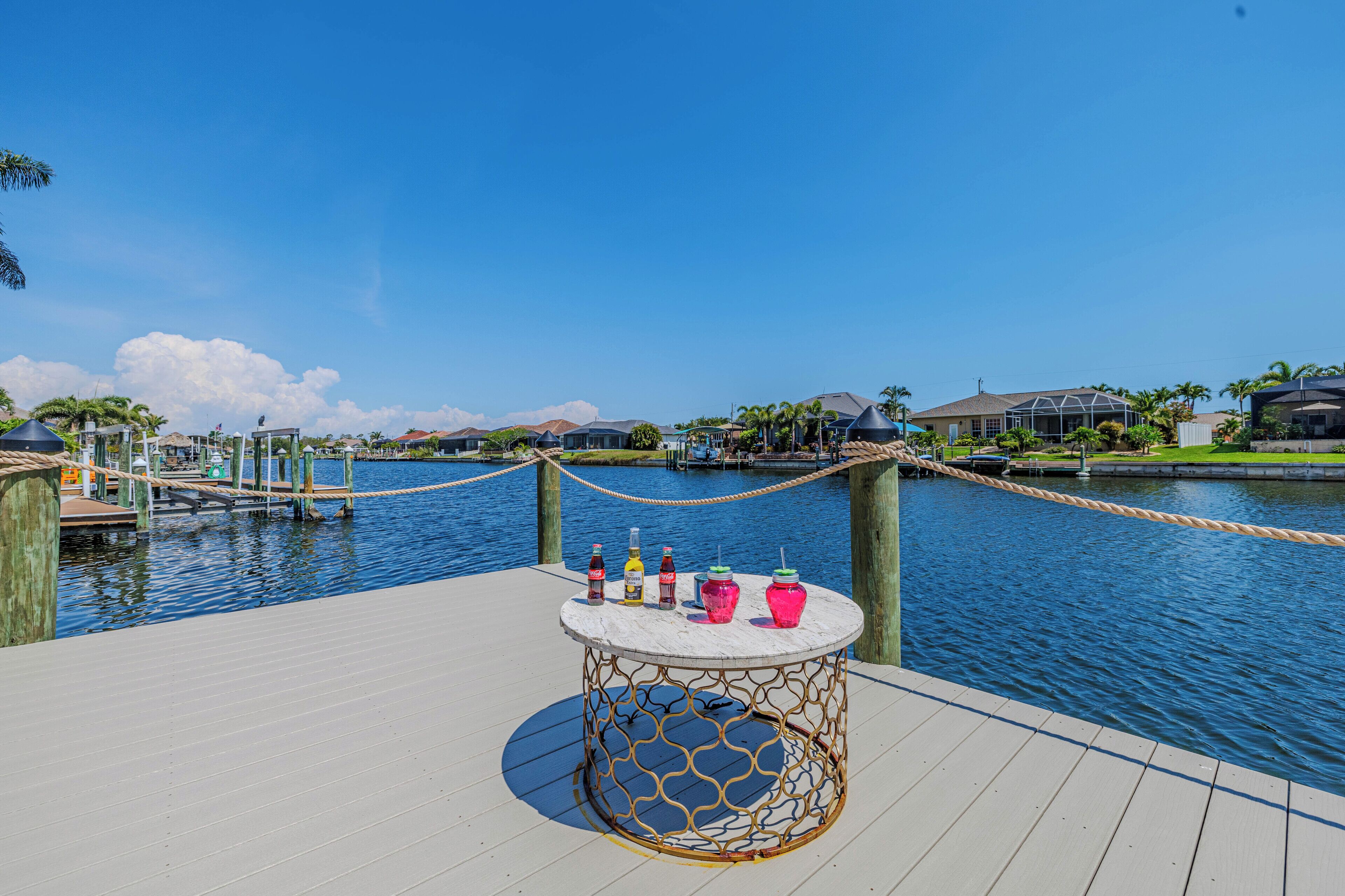 A dock with a rope barrier, a table with drinks, and a view of houses across the water.