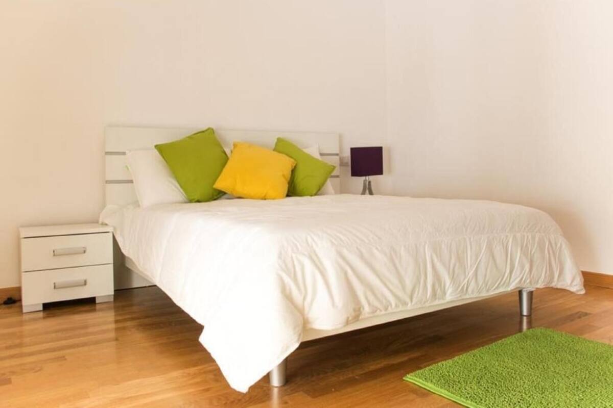 A neatly made bed with white bedding and colorful pillows, a small bedside table with drawers, and a green rug on a wooden floor.