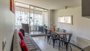 A modern living room with a sofa, a dining table with chairs, and a sliding glass door leading to a balcony. - Kennedy Apartments Los Militares (Santiago)