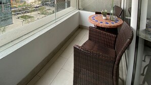 A balcony with a wicker chair, a small table with a vase, and a view of modern buildings. - Kennedy Apartments Los Militares (Santiago)
