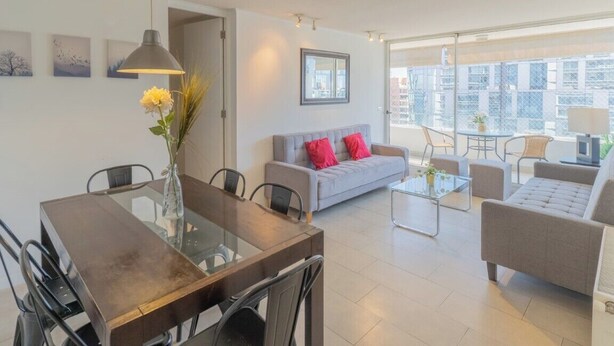 A modern living room with a dining area, grey sofa with red cushions, and a large window offering a city view. - Kennedy Apartments Los Militares (Santiago)