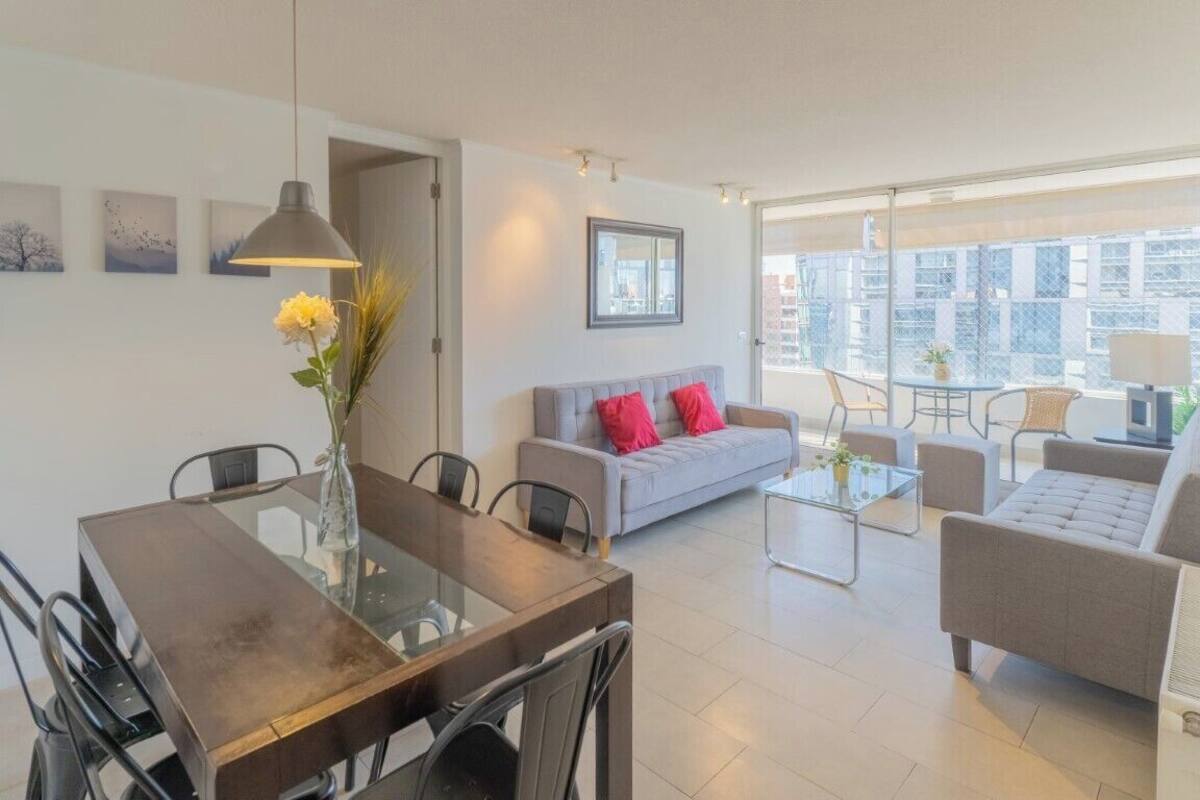 A modern living room with a dining area, grey sofa with red cushions, and a large window offering a city view.
