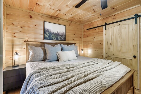 A wooden cabin bedroom with a bed, pillows, a nightstand, a wall-mounted picture, and a sliding door.
