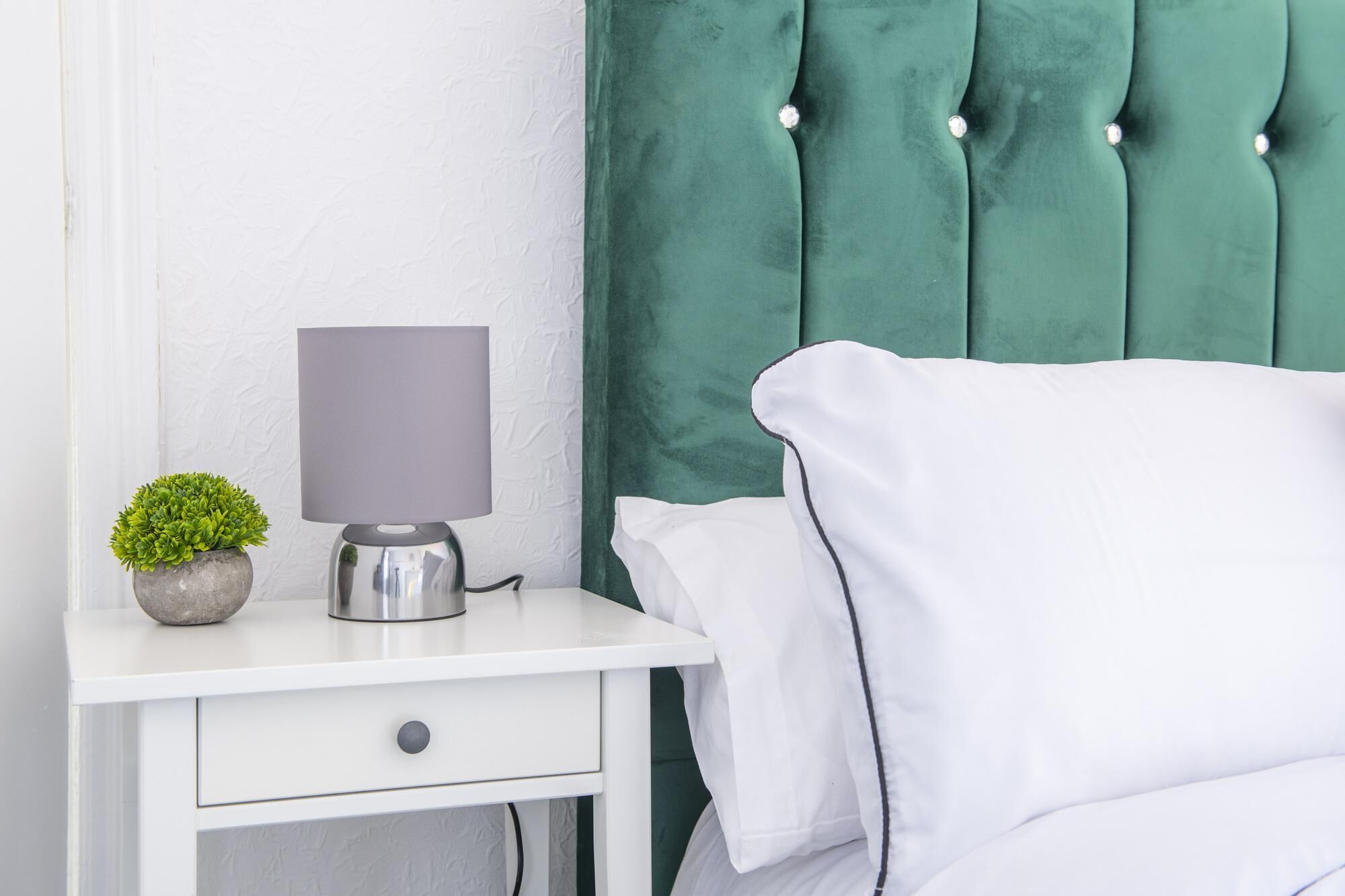 A neatly made bed with white linens, a small potted plant, and a bedside lamp on a white nightstand.