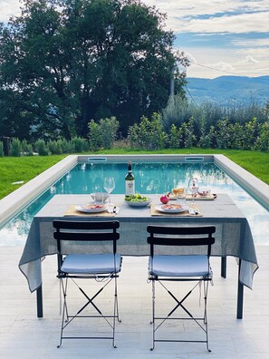 An outdoor dining setup by a pool with a table set for two, surrounded by greenery and a view of hills. - Villa Angela Ranza 55 (città di castello)