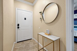 A hallway with a white door, a round mirror, a small table with a block of text, and a blue bed in the background. - Spacious 4 Bedroom Condo Resort Close to Disney (Davenport)