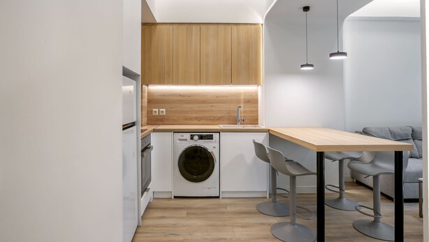 A compact kitchen with a washing machine, wooden cabinets, and a dining area with bar stools. - Porto Pireo by Superhost365 (Piraeus)