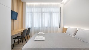 A modern hotel room with a large bed, a desk with chairs, and a large window with sheer curtains. - Porto Pireo by Superhost365 (Piraeus)