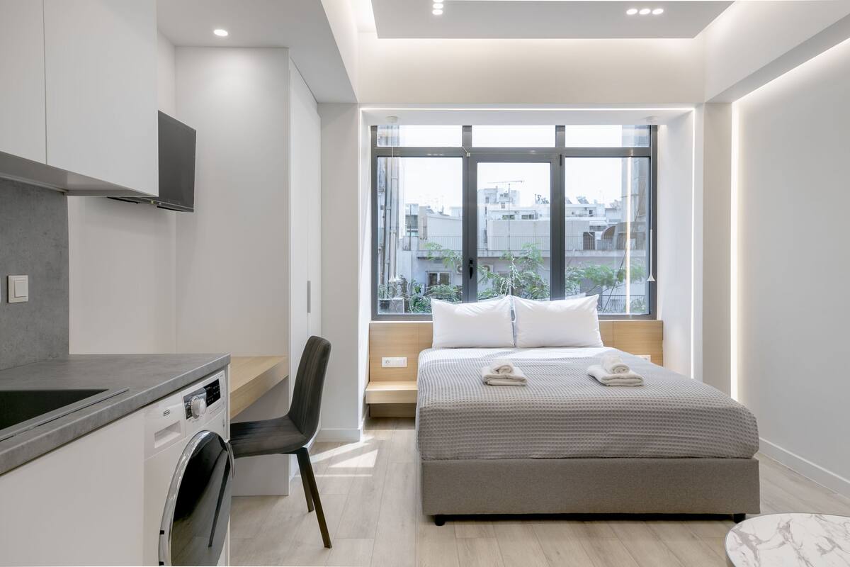 Une chambre d’hôtel moderne dotée d’une grande fenêtre, d’un lit, d’un lave-linge intégré et d’un coin salon.