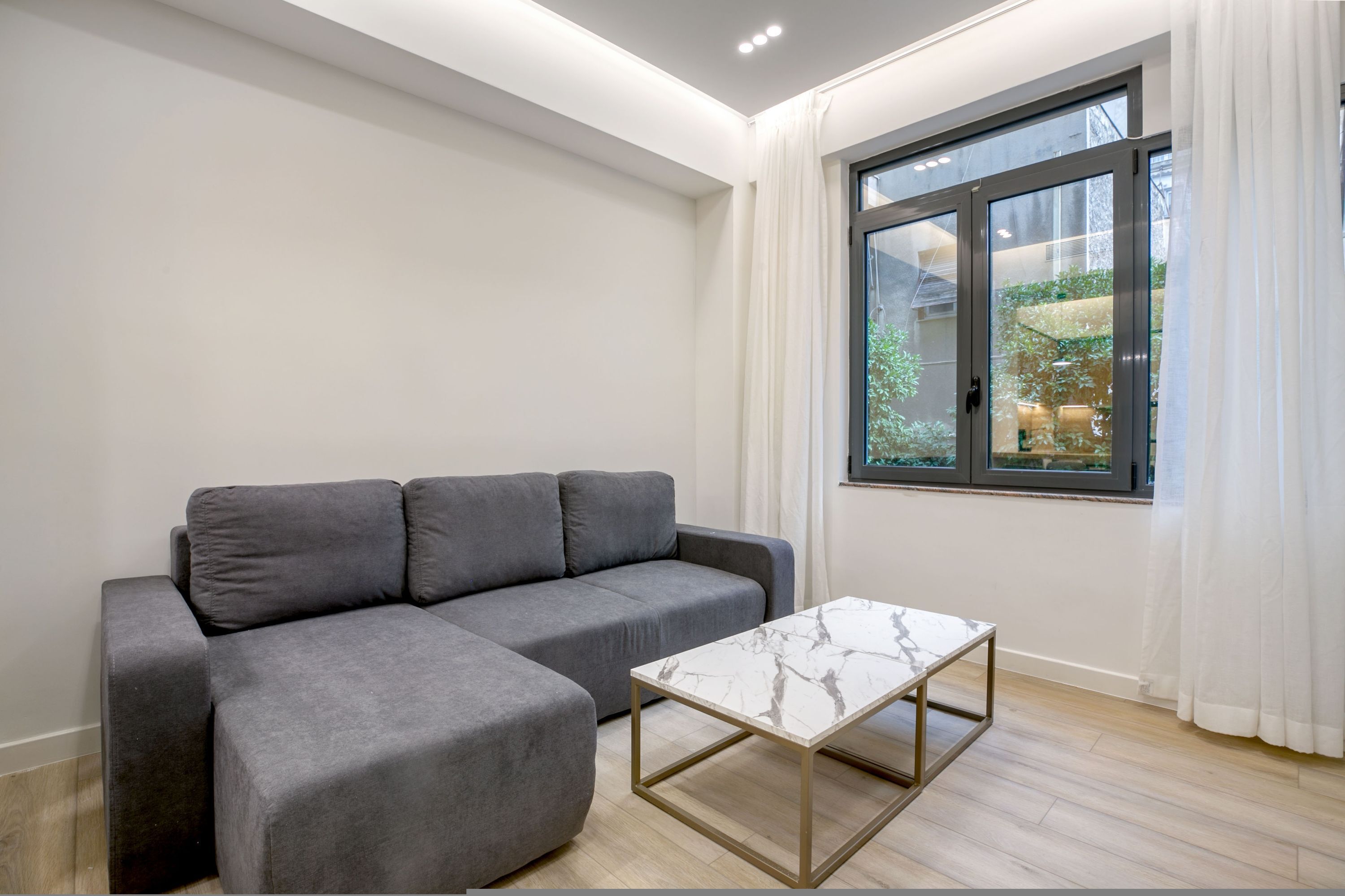 Een moderne woonkamer met een grijze modulaire bank, een marmeren salontafel en een groot raam met witte gordijnen.