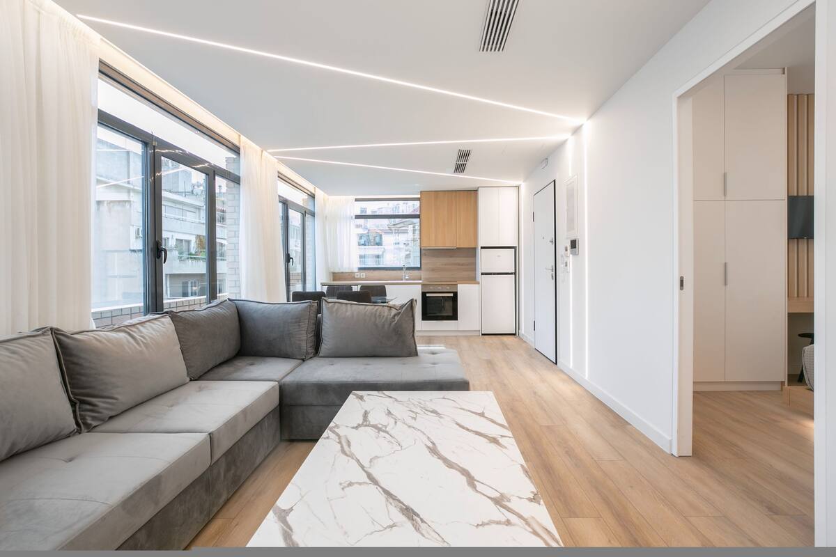 Un appartement moderne avec un espace de vie comprenant un canapé d’angle, une table basse en marbre et une cuisine en arrière-plan.