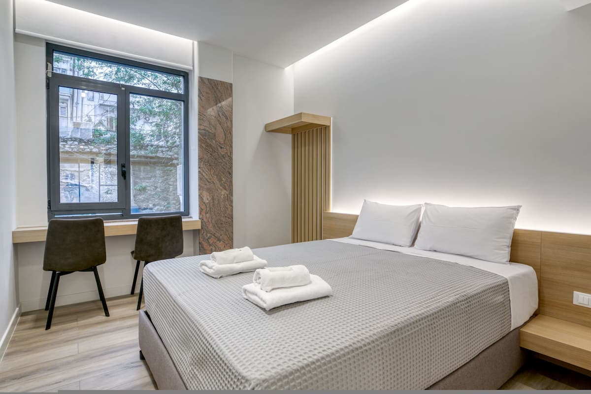 Une chambre d’hôtel moderne avec un grand lit, deux fauteuils, une fenêtre et une table de chevet en bois.