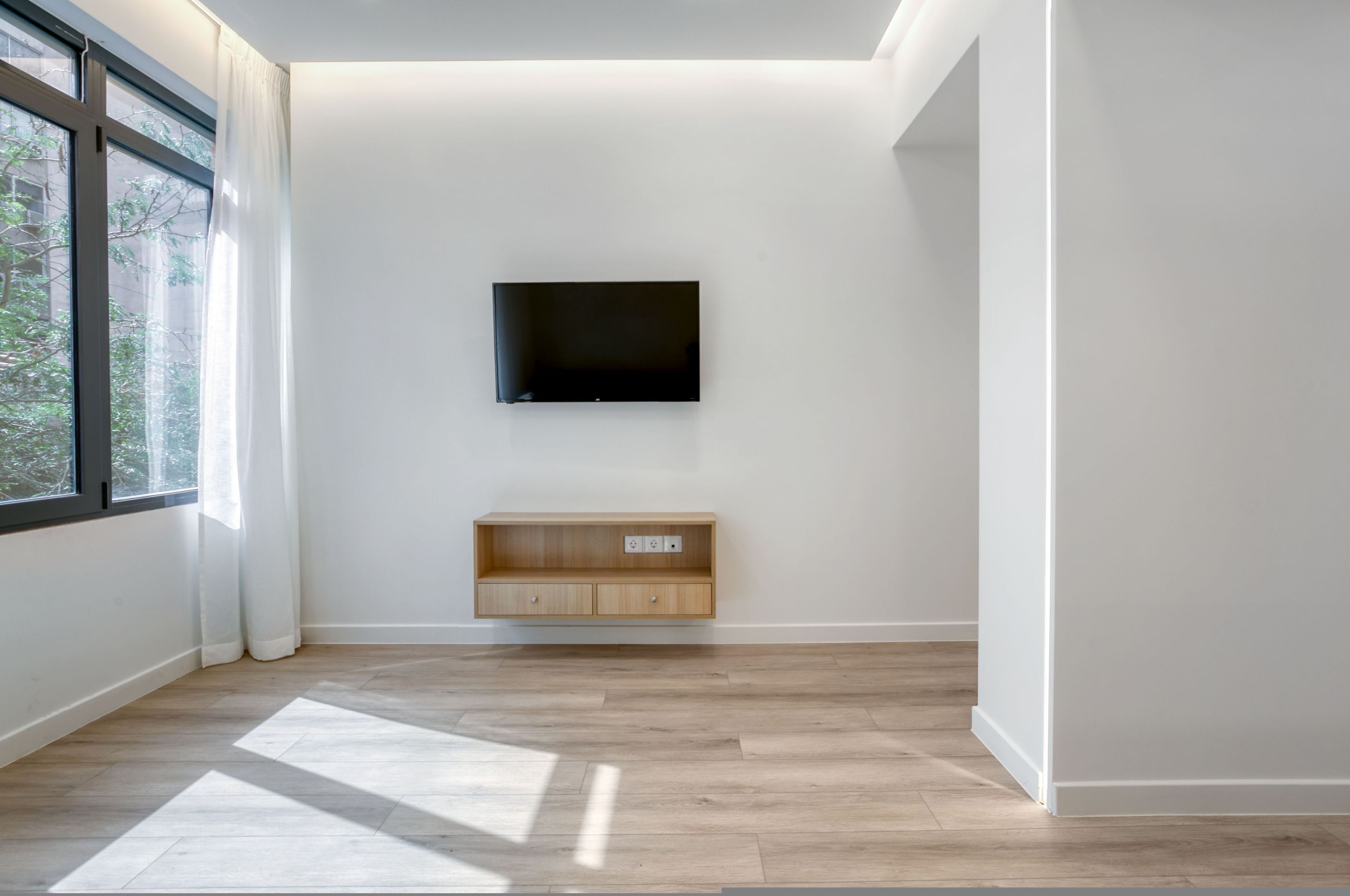 A modern room with a large window, a mounted TV, and a wooden shelf unit.