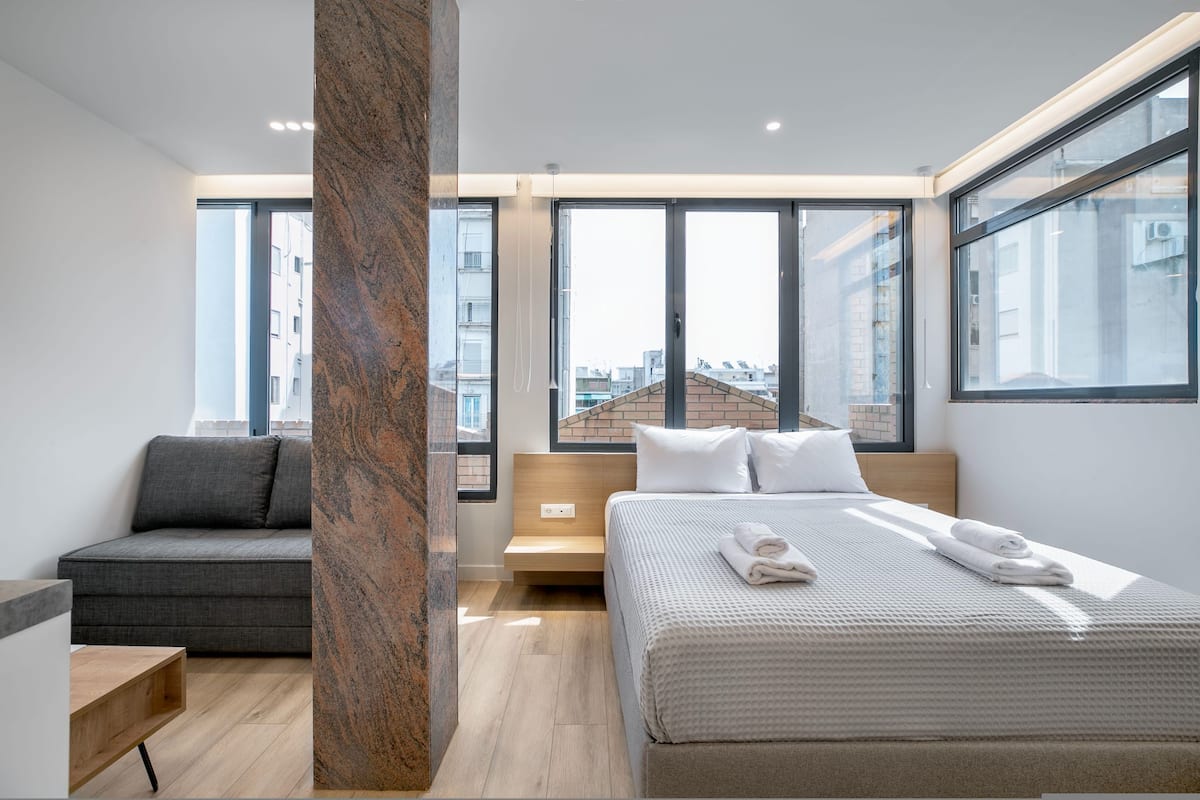 Une chambre d’hôtel moderne, dotée d’un grand lit, d’un canapé et offrant une vue sur la ville grâce à de grandes fenêtres.