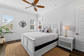 A bedroom with a white bed, a ceiling fan, a bedside table with a lamp, and a potted plant.