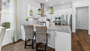 Una cocina con gabinetes blancos, encimera de granito, dos taburetes de mimbre, refrigerador de acero inoxidable y luces colgantes.