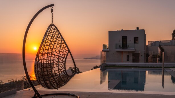 A modern outdoor seating area with a hanging chair by a pool, overlooking a body of water and a modern building. - Luxury Villa Verde (Malevizi)