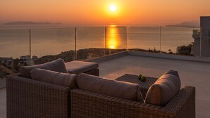 A balcony with wicker furniture and a glass railing overlooking a sunset over water. - Luxury Villa Verde (Malevizi)