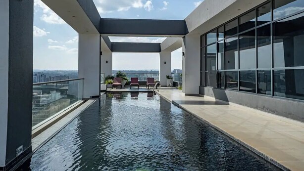 A rooftop infinity pool with a glass balustrade, red lounge chairs, and a view of the cityscape.