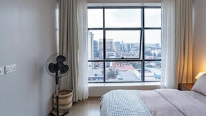 A bedroom with a large window, a standing fan, a bedside lamp, and a bed with a patterned bedspread. - Elite - 1 bedroom - pool & Gym

 (Nairobi)
