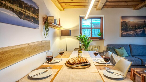 A dining area with a wooden table set for four, two wine glasses, a loaf of bread, and a bottle of wine.