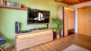 A modern living room with a large flat-screen TV mounted on the wall, wooden furniture, a bookshelf, and a potted plant.
